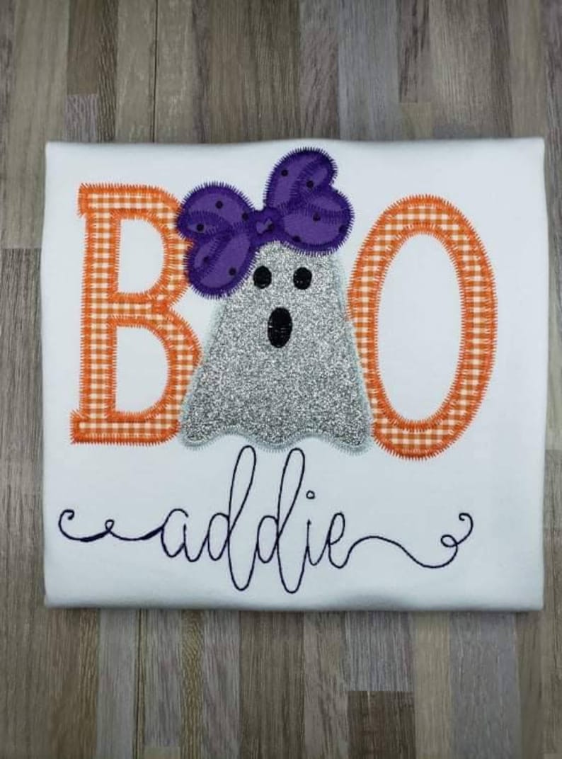 May include: White t-shirt with an embroidered design of the word "BOO" with a ghost wearing a purple bow. The name "addie" is embroidered below the design.