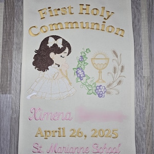 May include: A white banner with gold embroidery that reads "First Holy Communion". A girl with brown hair and a white dress is praying. A chalice with grapes and wheat is embroidered on the right side of the banner. The banner also has pink embroidery that reads "Ximena April 26, 2025 St. Marianne School."