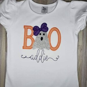 May include: White t-shirt with an orange and white checkered "BOO" with a ghost wearing a purple bow. The text "addie" is written in cursive below the ghost.
