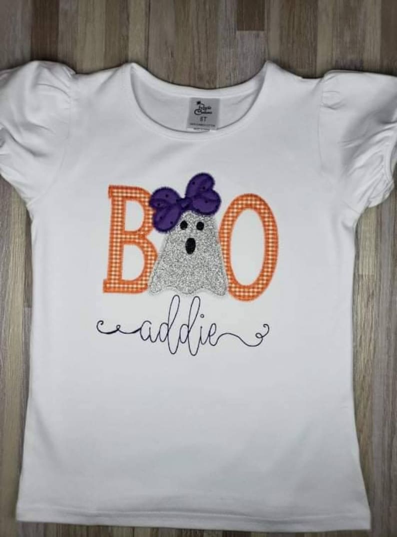 May include: White t-shirt with an orange and white checkered "BOO" with a ghost wearing a purple bow. The text "addie" is written in cursive below the ghost.