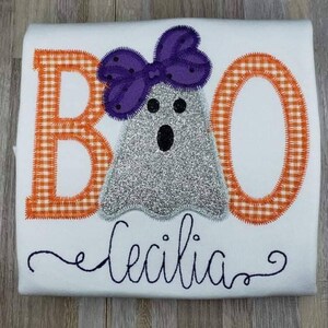 May include: A white t-shirt with an embroidered design of a ghost wearing a purple bow. The ghost is silver and glittery. The letters "BOO" are embroidered in orange and white gingham. The name "Cecilia" is embroidered in black cursive below the ghost.