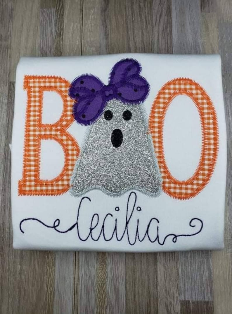 May include: A white t-shirt with an embroidered design of a ghost wearing a purple bow. The ghost is silver and glittery. The letters "BOO" are embroidered in orange and white gingham. The name "Cecilia" is embroidered in black cursive below the ghost.