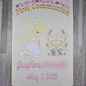 May include: A white fabric banner with pink hearts and gold embroidery. The banner reads "First Communion" and features a girl kneeling with a chalice and grapes. The banner is personalized with the name "Josephine" and the date "May 3, 2025".