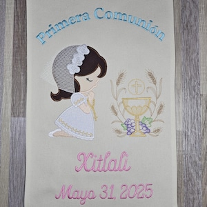 May include: A beige fabric banner with embroidered text that reads "Primera Comunión" in blue. The banner also features an embroidered image of a girl in a white dress kneeling with a chalice and wheat stalks. The text "Xitlalí Mayo 31, 2025" is embroidered in pink at the bottom.