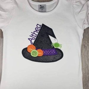 K&ouml;nnte beinhalten: Wei&szlig;es T-Shirt mit einem Hexenhut-Design. Der schwarze Hut hat ein lila Band mit Pailletten, orangefarbenen und gr&uuml;nen Akzenten und eine gr&uuml;ne Spinne. Der Name "Althea" ist lila gestickt. Das Shirt hat kurze, gepuffte &Auml;rmel.
