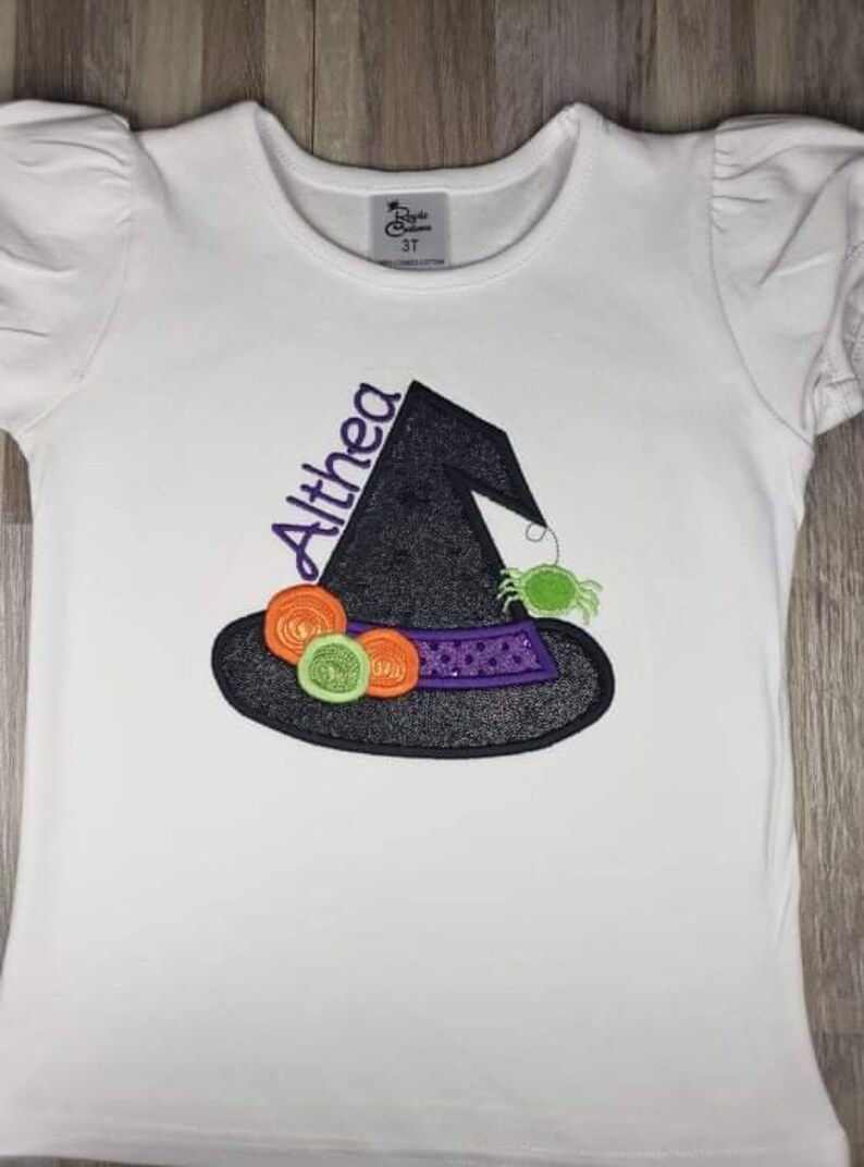 K&ouml;nnte beinhalten: Wei&szlig;es T-Shirt mit einem Hexenhut-Design. Der schwarze Hut hat ein lila Band mit Pailletten, orangefarbenen und gr&uuml;nen Akzenten und eine gr&uuml;ne Spinne. Der Name "Althea" ist lila gestickt. Das Shirt hat kurze, gepuffte &Auml;rmel.