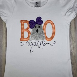 May include: White short-sleeved shirt with a ghost wearing a purple bow and the letters "BOO" in orange and white gingham. The name "rayanne" is written in cursive below the ghost.