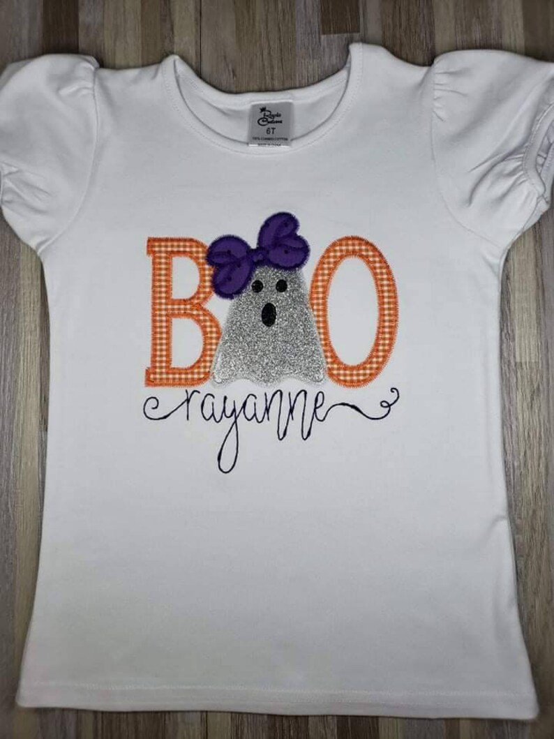 May include: White short-sleeved shirt with a ghost wearing a purple bow and the letters "BOO" in orange and white gingham. The name "rayanne" is written in cursive below the ghost.