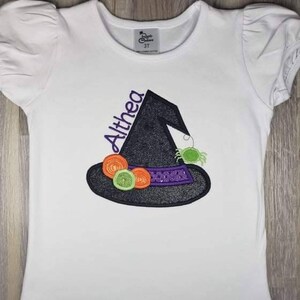 K&ouml;nnte beinhalten: Wei&szlig;es Kurzarm-T-Shirt mit einem schwarzen Hexenhut-Design und dem Namen "Althea" in Lila. Der Hut ist mit orangefarbenen und gr&uuml;nen Kreisen, einem lila Band und einer gr&uuml;nen Spinne verziert. Das Shirt ist in Gr&ouml;&szlig;e 3T.