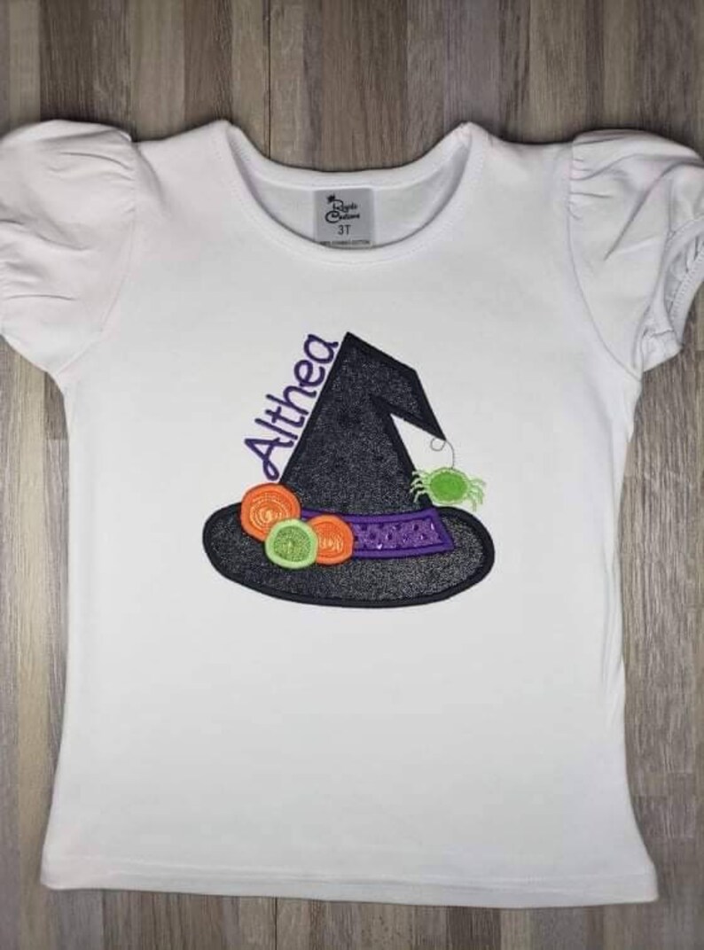 K&ouml;nnte beinhalten: Wei&szlig;es Kurzarm-T-Shirt mit einem schwarzen Hexenhut-Design und dem Namen "Althea" in Lila. Der Hut ist mit orangefarbenen und gr&uuml;nen Kreisen, einem lila Band und einer gr&uuml;nen Spinne verziert. Das Shirt ist in Gr&ouml;&szlig;e 3T.