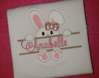 May include: White fabric with an embroidered bunny design. The bunny has pink ears, a bow, and paws. The name "Anabelle" is embroidered in pink script. The background is a sparkly pink.