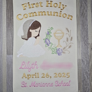 May include: A white banner with gold embroidery that reads "First Holy Communion". The banner features an embroidered image of a girl in a white dress and veil, holding a prayer book. The banner also includes a chalice and grapes. The text "Lilyth April 26, 2025 St. Marianne School" is embroidered in pink on the banner.