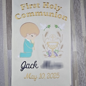 May include: A white fabric banner with gold embroidery that says "First Holy Communion". There is a small boy kneeling in prayer with a chalice and host, wheat, and doves embroidered on the banner. The boy's name, "Jack", and the date "May 10, 2025" are also embroidered on the banner.