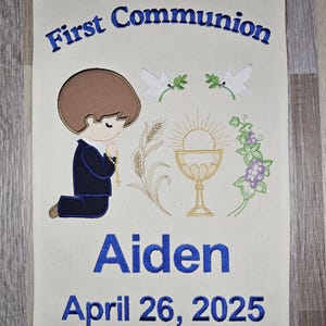 May include: A white fabric banner with blue embroidery. The banner says "First Communion" at the top, and "Aiden April 26, 2025" at the bottom. The banner features an embroidered image of a boy kneeling in prayer, a chalice, doves, and wheat.