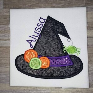 K&ouml;nnte beinhalten: Wei&szlig;er Stoff mit einem gestickten Hexenhut-Design. Der schwarze Hut hat ein lila Band, orangefarbene und gr&uuml;ne Verzierungen und eine gr&uuml;ne Spinne. Der Name "Alyssa" ist in Lila auf dem Hut gestickt.
