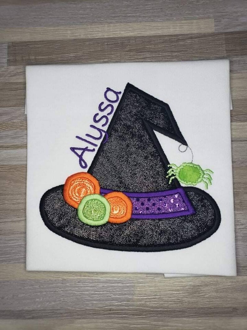 K&ouml;nnte beinhalten: Wei&szlig;er Stoff mit einem gestickten Hexenhut-Design. Der schwarze Hut hat ein lila Band, orangefarbene und gr&uuml;ne Verzierungen und eine gr&uuml;ne Spinne. Der Name "Alyssa" ist in Lila auf dem Hut gestickt.