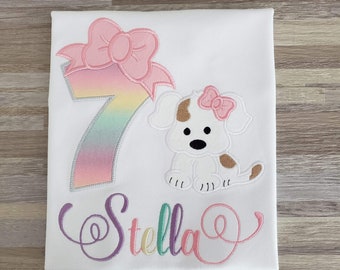 Puede incluir: Una camiseta blanca con un número 7 de colores arcoíris, un lazo rosa y un cachorro blanco con manchas marrones que lleva un lazo rosa. El nombre "Stella" está escrito en colores arcoíris debajo del cachorro.