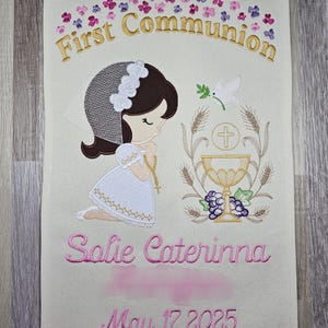 May include: A white banner with embroidered text that reads "First Communion" in gold thread. The banner features a girl in a white dress and veil, holding a rosary. The text "Solie Caterinna" and "May 17, 2025" are embroidered in pink thread.
