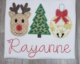 May include: White t-shirt with a red reindeer, green Christmas tree, and gold ornament with a red bow. The name "Rayanne" is embroidered in red below the ornaments.