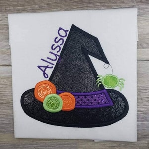 K&ouml;nnte beinhalten: Wei&szlig;er Stoff mit einem Design eines schwarzen Hexenhuts. Der Hut hat ein lilafarbenes Band mit Pailletten und ist mit orangefarbenen und gr&uuml;nen Kreisen verziert. Eine gr&uuml;ne Spinne h&auml;ngt am Hut, und der Name "Alyssa" ist lila gestickt.