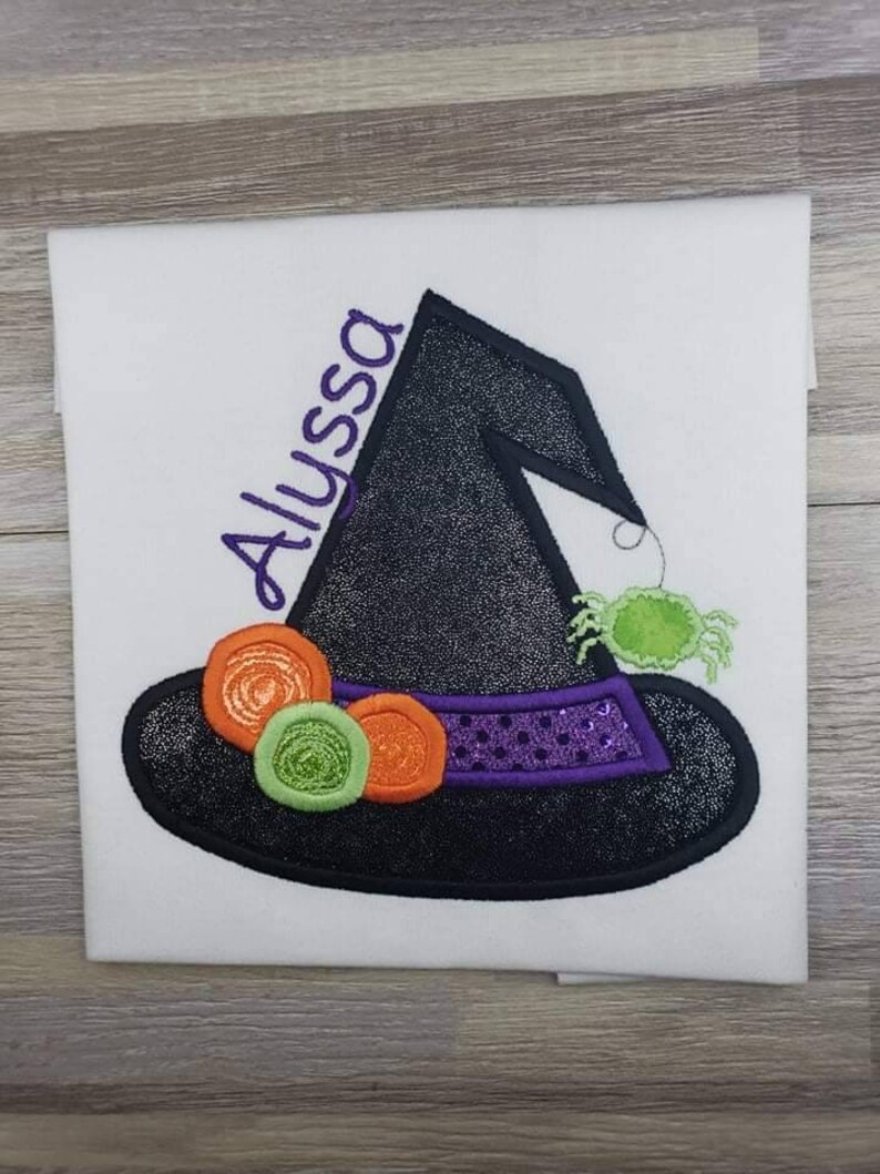 K&ouml;nnte beinhalten: Wei&szlig;er Stoff mit einem Design eines schwarzen Hexenhuts. Der Hut hat ein lilafarbenes Band mit Pailletten und ist mit orangefarbenen und gr&uuml;nen Kreisen verziert. Eine gr&uuml;ne Spinne h&auml;ngt am Hut, und der Name "Alyssa" ist lila gestickt.