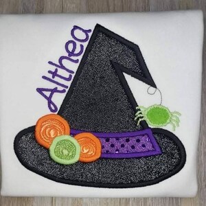 K&ouml;nnte beinhalten: Wei&szlig;es T-Shirt mit einem schwarzen Hexenhut-Design. Der Hut hat ein lila Band mit Pailletten, orangefarbenen und gr&uuml;nen Akzenten und eine gr&uuml;ne Spinne. Der Name "Althea" ist in Lila gestickt.