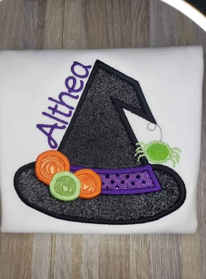 K&ouml;nnte beinhalten: Wei&szlig;es T-Shirt mit einem schwarzen Hexenhut-Design. Der Hut hat ein lila Band mit Pailletten, orangefarbenen und gr&uuml;nen Akzenten und eine gr&uuml;ne Spinne. Der Name "Althea" ist in Lila gestickt.