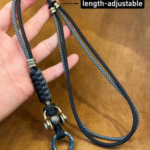 Könnte beinhalten: Verstellbares schwarzes und graues Schlüsselband mit Metallring und dekorativen Elementen. Das Schlüsselband hat ein geflochtenes Design und den Text "length-adjustable". Es ist für Schlüssel oder kleine Gegenstände konzipiert.