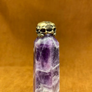 Könnte beinhalten: Eine violette Amethyst-Kristallspitze mit einem Messing-Schädel-Topper. Der Schädel ist mit drei Gesichtern verziert.