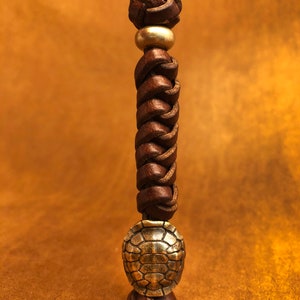 May include: A brown leather cord with a braided design and two metal accents. The top accent is a round bead and the bottom accent is a turtle charm.