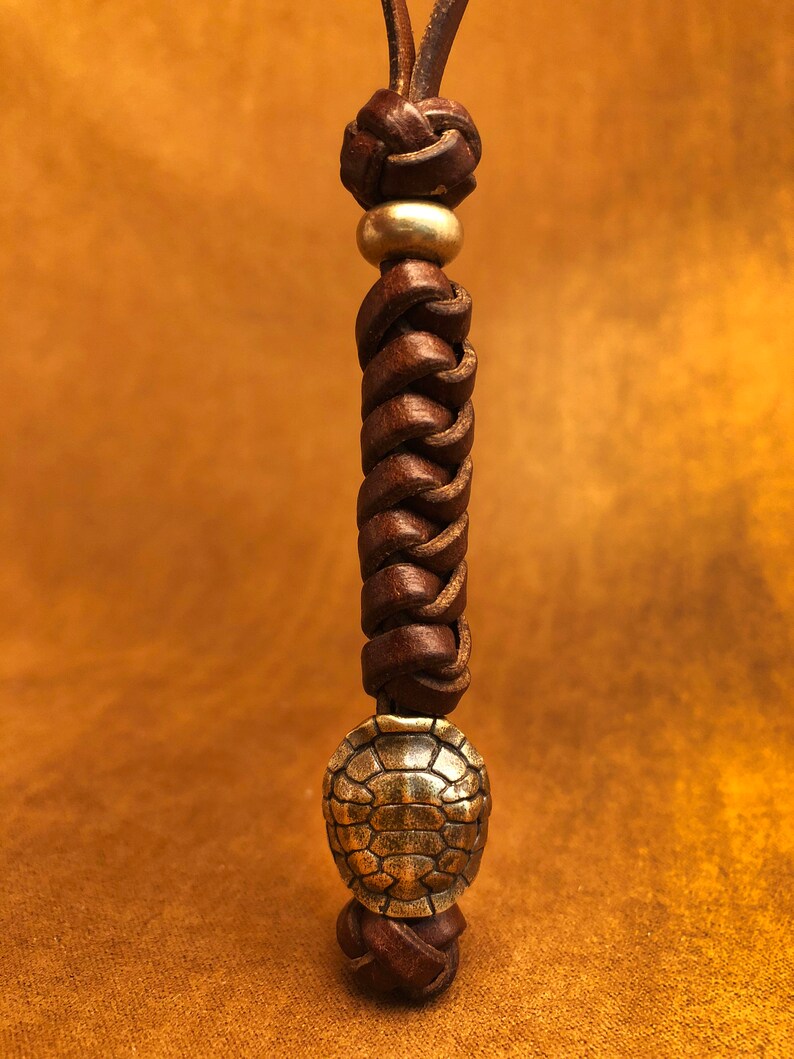 May include: A brown leather cord with a braided design and two metal accents. The top accent is a round bead and the bottom accent is a turtle charm.
