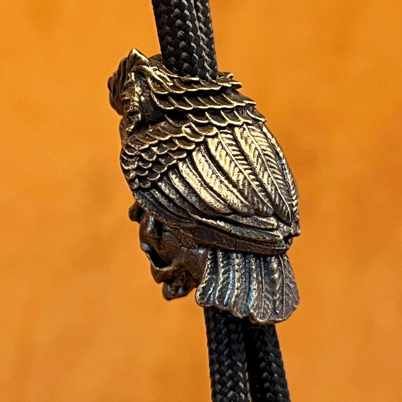 Puede incluir: Una corbata bolo de color bronce con un dise&ntilde;o detallado de b&uacute;ho. El b&uacute;ho tiene intrincados detalles de plumas y est&aacute; sujeto a un cord&oacute;n trenzado negro. La cabeza del b&uacute;ho tiene una apariencia de calavera. El fondo es de color naranja liso.