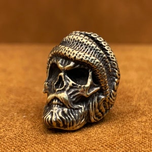 May include: A bronze-coloured skull sculpture wearing a knitted cap. The skull has a textured surface with visible facial features, including eye sockets, a nose, and a beard. The cap has a ribbed pattern.