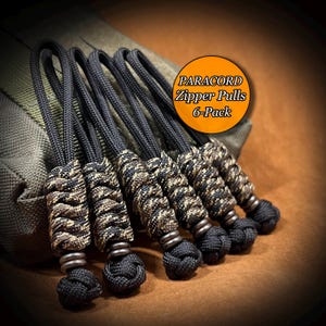 May include: A set of six black and camouflage paracord zipper pulls, each featuring a braided cord loop, a textured knot section, and a solid black knot at the base. An orange circular graphic displays the text PARACORD Zipper Pulls 6-Pack.