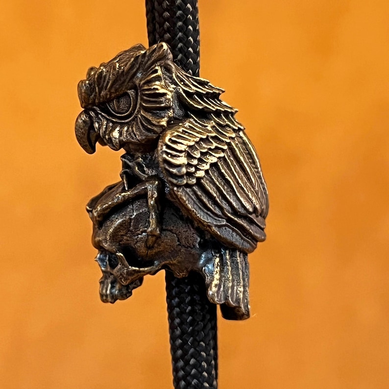 Puede incluir: Un colgante de b&uacute;ho de color bronce con detalles intrincados, posado sobre una calavera. El colgante est&aacute; suspendido de un cord&oacute;n negro trenzado. Las plumas del b&uacute;ho y los rasgos de la calavera est&aacute;n claramente definidos.