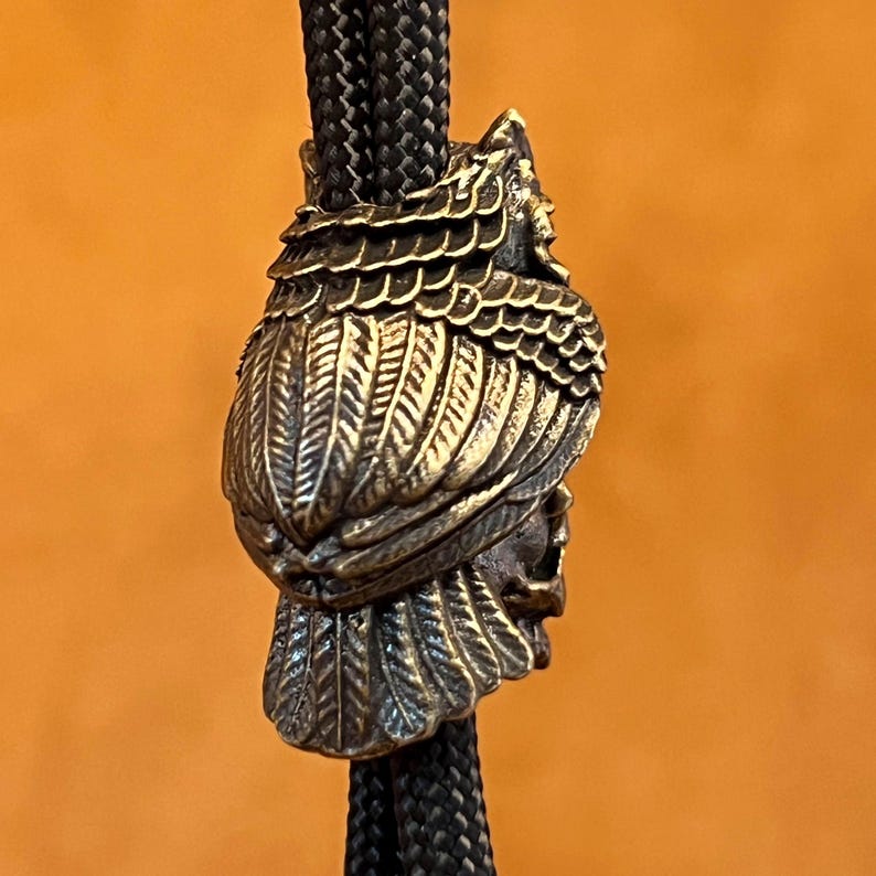 Puede incluir: Colgante de corbata bolo de color bronce con un dise&ntilde;o detallado de un jefe nativo americano con tocado de plumas. El colgante est&aacute; unido a un cord&oacute;n trenzado negro, sobre un fondo naranja.