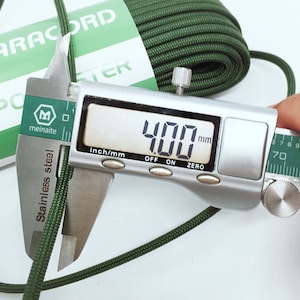 May include: A coil of olive green paracord with a white paper label reading PARACORD and POLYESTER. A stainless steel digital caliper is measuring the diameter of the cord, showing a clear reading of 4.00 mm on the digital display screen.