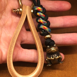 May include: A brass key chain with a leather loop and a frog charm. The key chain has a black and colorful braided cord with a black bead.