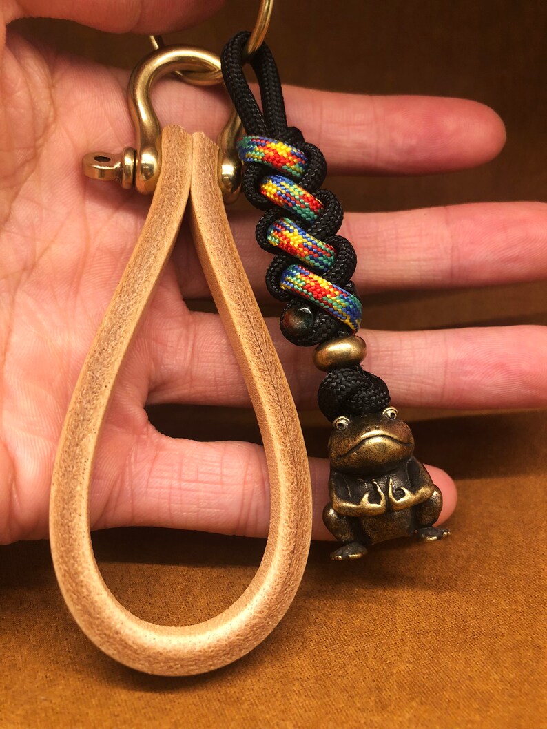 May include: A brass key chain with a leather loop and a frog charm. The key chain has a black and colorful braided cord with a black bead.