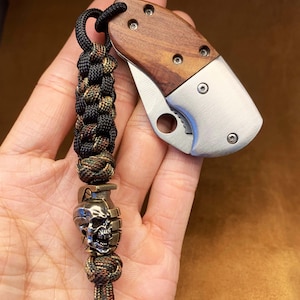 May include: A small, silver pocket knife with a wooden handle and a skull-shaped bead on a black and green paracord lanyard.