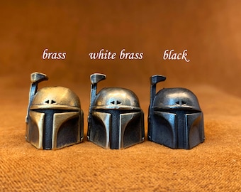 Op de afbeelding: Drie Boba Fett helm-vormige sleutelhangers in messing, wit messing en zwart. De sleutelhangers zijn gerangschikt van links naar rechts in de volgorde van hun afwerkingen.