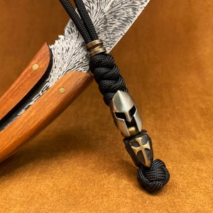 Könnte beinhalten: Ein schwarzes Paracord-Lanyard mit einem silbernen und goldenen Metallanhänger mit einem Helm und einem Kreuz. Das Lanyard ist an einem Messer mit einem Holzgriff und einer gemusterten Klinge befestigt.