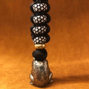 May include: A silver frog charm with a detailed design hangs from a black and white paracord lanyard with a brass bead.