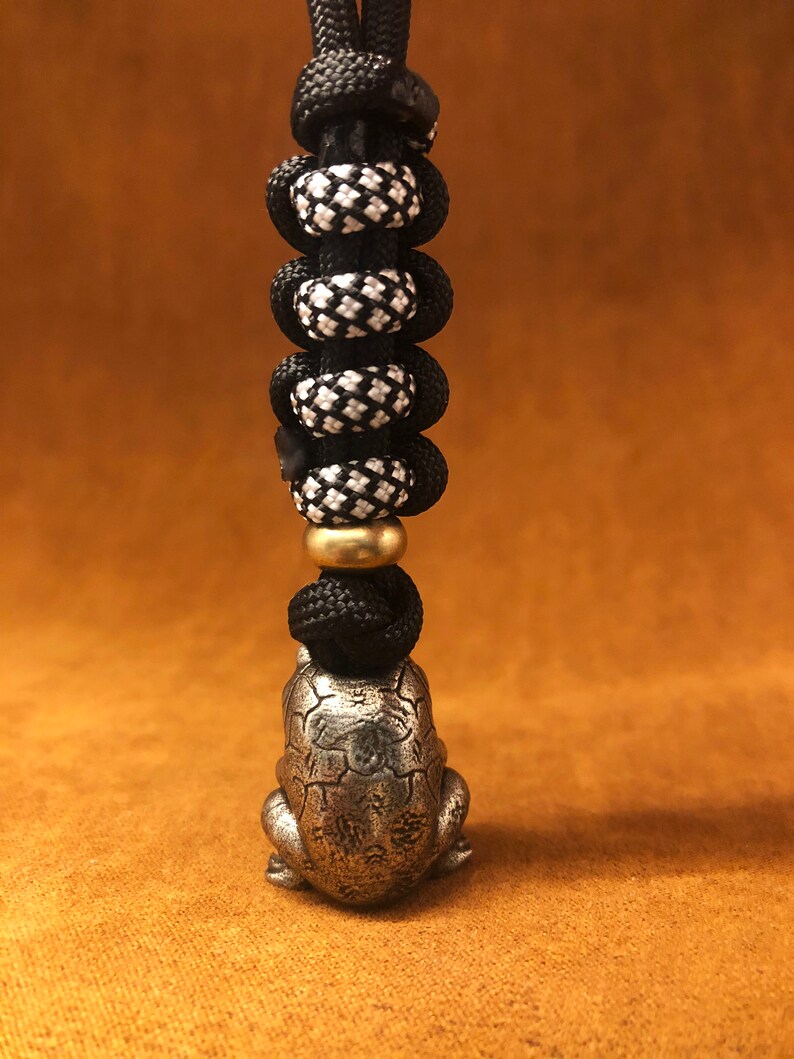 May include: A silver frog charm with a detailed design hangs from a black and white paracord lanyard with a brass bead.