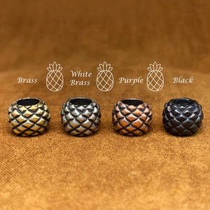 May include: Four pineapple-shaped beads in different finishes: brass, white brass, purple, and black. The beads are all the same size and shape, and they are arranged in a row from left to right.