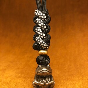 May include: Black and white checkered paracord lanyard with a black bead and a brass frog charm. The lanyard has a loop at the top for attaching to a keychain or bag.