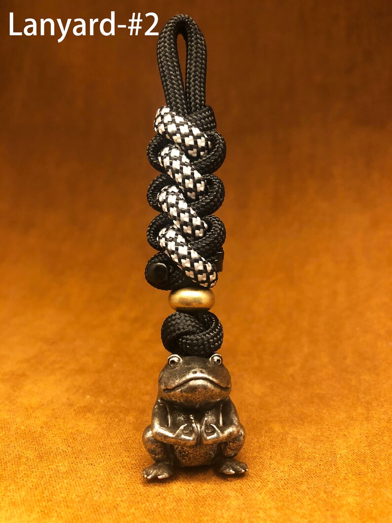 May include: Black and white checkered paracord lanyard with a black bead and a brass frog charm. The lanyard has a loop at the top for attaching to a keychain or bag.