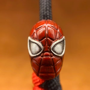May include: A red and white Spider-Man mask charm with a detailed design. The charm is attached to a black cord.