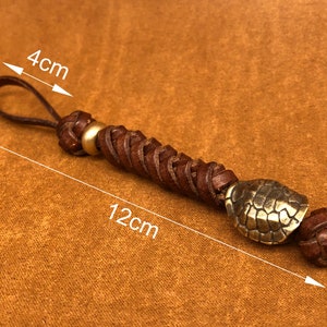 May include: A brown leather keychain with a braided design and a gold-toned turtle charm. The keychain measures 12 cm in length and 4 cm in width.