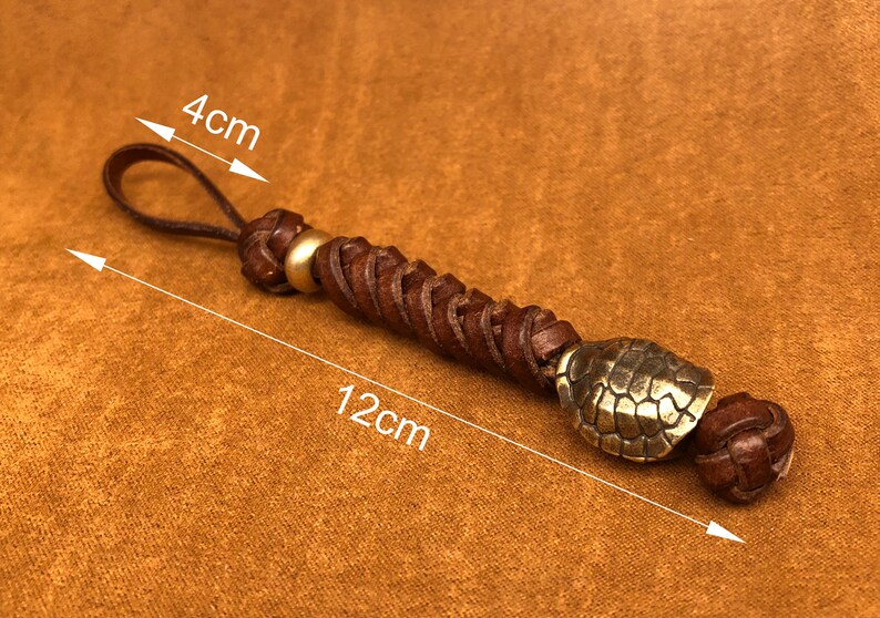 May include: A brown leather keychain with a braided design and a gold-toned turtle charm. The keychain measures 12 cm in length and 4 cm in width.